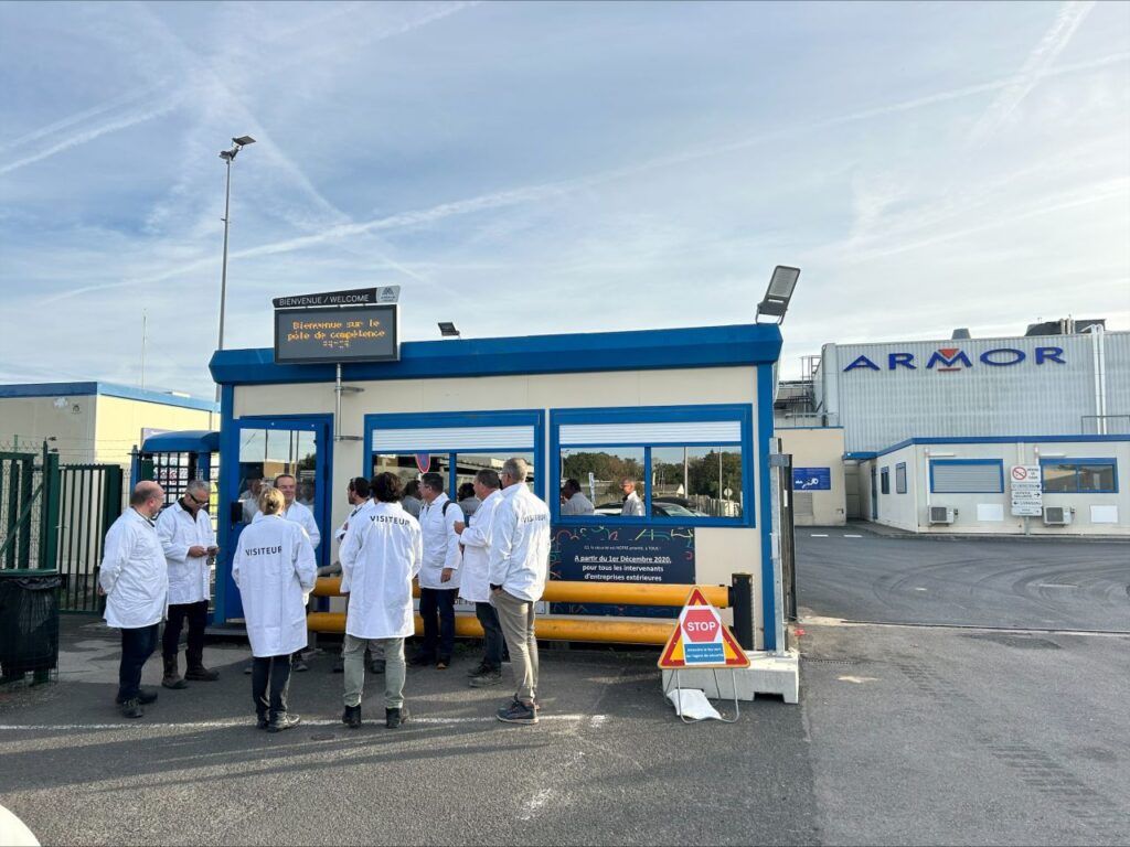 Photo de groupe de visiteurs en blouses blanches devant l’entrée du site ARMOR-IIMAK, avec la signalétique du site visible en arrière-plan.