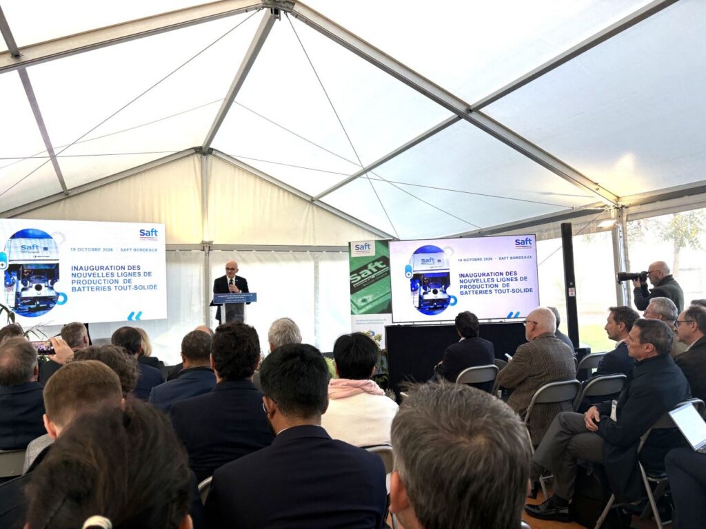 Vue d’ensemble de la cérémonie, avec les invités assis face à un intervenant au pupitre. Deux écrans affichent l’annonce de l’inauguration des nouvelles lignes de production de batteries tout-solide, organisée le 19 octobre 2026 à Bordeaux. À droite, un photographe immortalise la prise de parole.